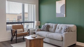 A living room with a sofa, armchair, and coffee table, a window with blinds, and a green wall with a framed picture. - Landing | Spectacular 1BD, Gym, Courtyard (Goodyear)