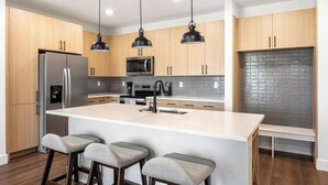 A modern kitchen with wooden cabinets, a stainless steel refrigerator, a built-in microwave, a large island with bar stools, and pendant lighting. - Landing | Spectacular 2BD, Game Room, Courtyard (Goodyear)