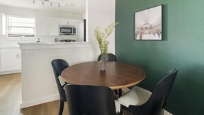 Et spiseområde med et rundt bord, fire stole, en vase med blomster, et køkken i baggrunden og et billede i ramme på væggen.