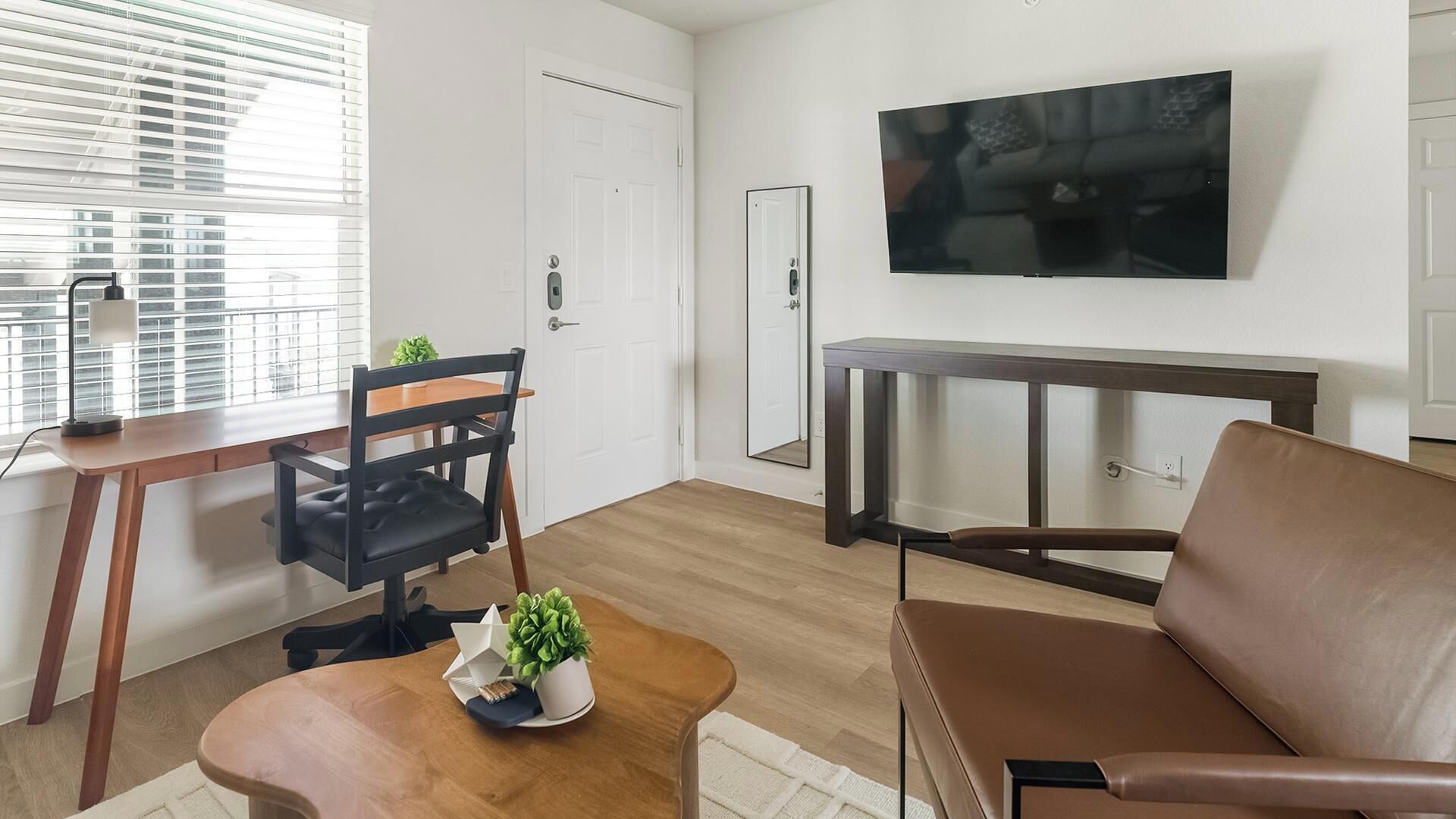 A modern living room with a wooden floor, a brown leather sofa, a dining table with chairs, a small coffee table with plants, and a flat-screen TV mounted on the wall.