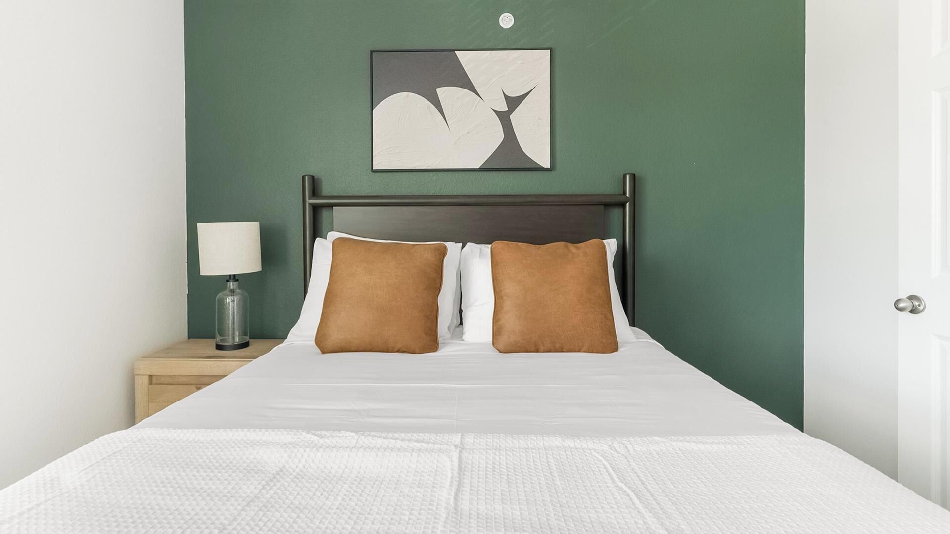 A neatly made bed with white bedding and two brown pillows, a bedside table with a lamp, and a wall-mounted abstract art piece.