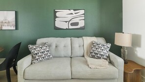 Ein helles Sofa mit zwei gemusterten Kissen, einer gestrickten Decke und einem gerahmten abstrakten Kunstwerk an einer grünen Wand.