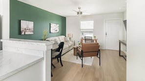 A modern living room with a green accent wall, a white sofa, a dining area with a table and chairs, and a ceiling fan. - Landing | Modern 2BD in Bear Creek (Houston)