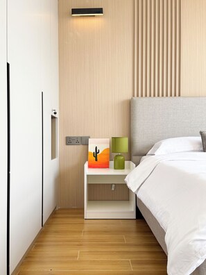 A modern bedroom with a wooden floor, a bed with white bedding, a nightstand with a cactus artwork, a green lamp, and a wall-mounted light fixture. - Plum Skyline Kilimani (Nairobi)