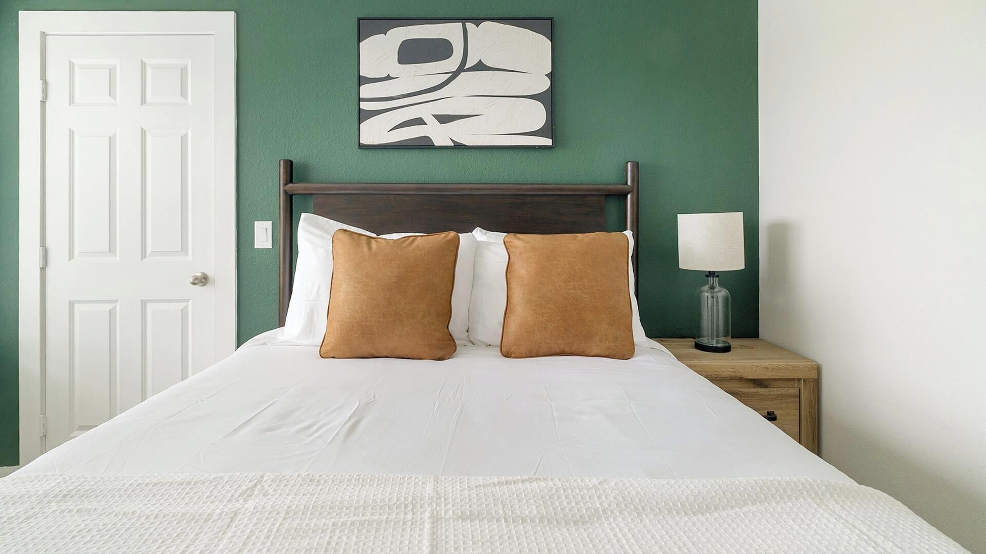A neatly made bed with white bedding and two brown pillows, a wooden headboard, a green wall with a framed abstract art piece, a white door, and a bedside table with a lamp.