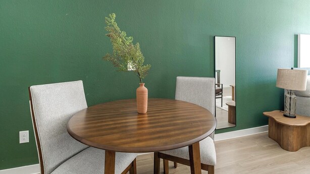 Une table ronde en bois, surmontée d’un vase texturé et d’une plante, est flanquée de deux chaises grises à revêtement en tissu. Le tout est placé devant un mur vert, où l’on aperçoit un miroir et une lampe posée sur une petite table d’appoint en bois.