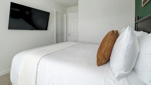 Habitació d'hotel amb llit, roba de llit blanca, almoïna marró, televisió penjada a la paret i una porta.
