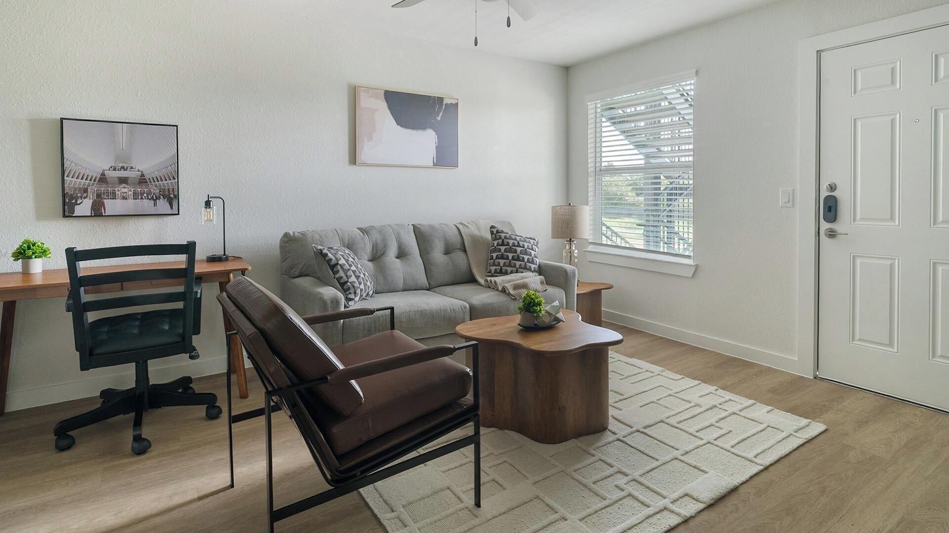 A modern living room with a sofa, a leather chair, a coffee table, a desk with a lamp, and a picture on the wall.