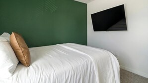En pænt redt seng med hvide sengetøj og en enkelt brun pude, en grøn væg og et sort tv ophængt på væggen.