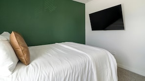 Ein ordentlich bezogenes Bett mit weißer Bettwäsche und einem einzelnen braunen Kopfkissen, eine grüne Wand und ein schwarzer Wandfernseher.