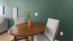 A round wooden table with a vase of green foliage, surrounded by grey upholstered chairs, against a teal wall with a white switchboard. - Landing | Chic 1BD in Bear Creek (Houston)