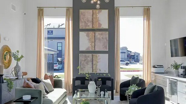 Un moderno salón con sofá, sillones, una mesa de centro y una televisión. Hay ventanas amplias con cortinas, una lámpara de araña y obras de arte en la pared.