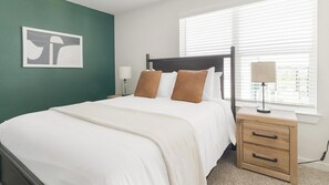A neatly made bed with white bedding and two brown pillows, a bedside table with a lamp, and a window with blinds. - Landing | Stunning 1BD in Bear Creek (Houston)