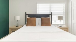 Uma cama bem arrumada com roupa de cama branca e duas almofadas marrons, uma mesinha de cabeceira com abajur e uma janela com persianas.