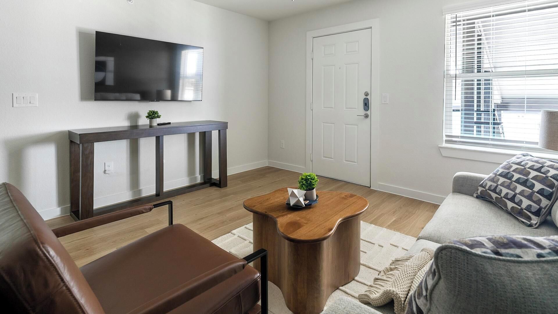 Ein Wohnzimmer mit einem braunen Ledersofa, einem Couchtisch aus Holz mit einer Pflanze, einem grauen Sofa mit gemusterten Kissen, einer weißen Tür und einem an der Wand befestigten Flachbildfernseher.