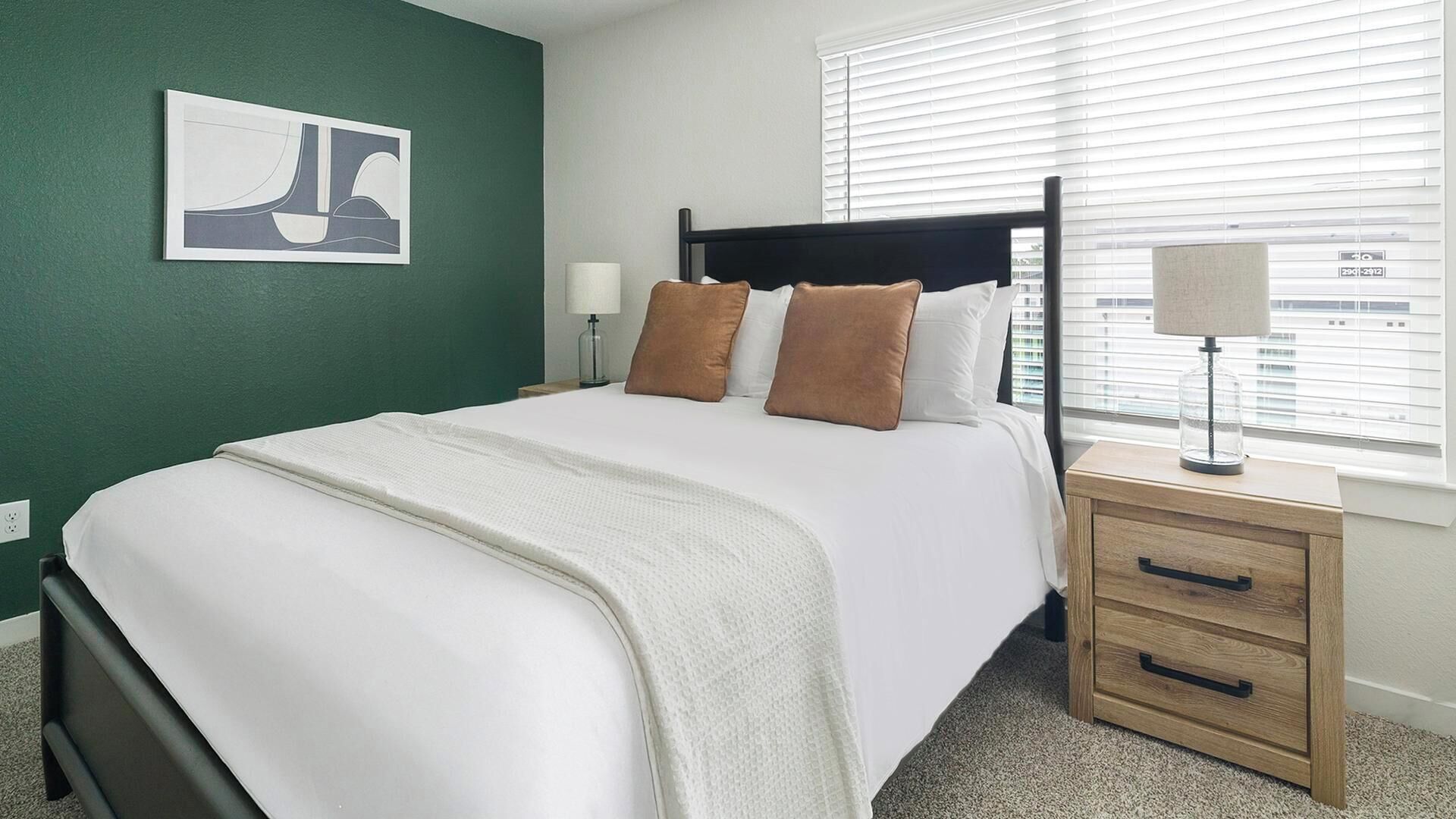 A neatly made bed with a white comforter and brown pillows, a bedside table with a lamp, and a window with blinds.