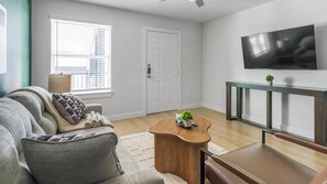 A living room with a sofa, a coffee table, a TV mounted on the wall, and a door. - Landing | Stylish 1BD in Bear Creek (Houston)