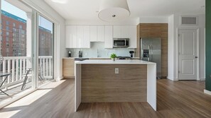 A modern kitchen with white cabinets, a central island, stainless steel appliances, and a view of a cityscape through large windows. - Landing | Stylish 2BD, Gym, Rooftop Lounge (Chevy Chase)