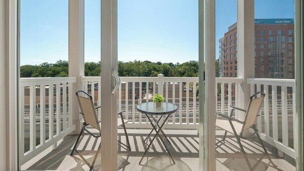 En balkon med et glasbord og to stole, et hvidt gelænder og udsigt til en bygning på den anden side af gaden.