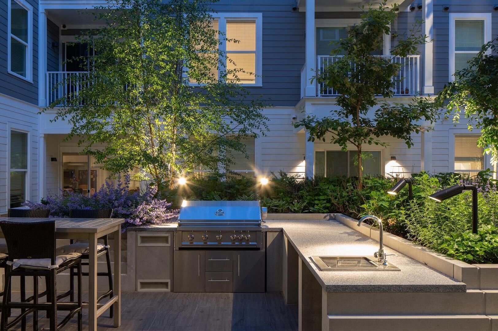 An outdoor kitchen area with a grill, dining table, and chairs, surrounded by greenery and a building in the background.