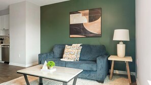 A living room with a sofa, a coffee table, a lamp, and a painting on the wall. - Landing | Spectacular 1BD, Gym, Rooftop Lounge (Chevy Chase)