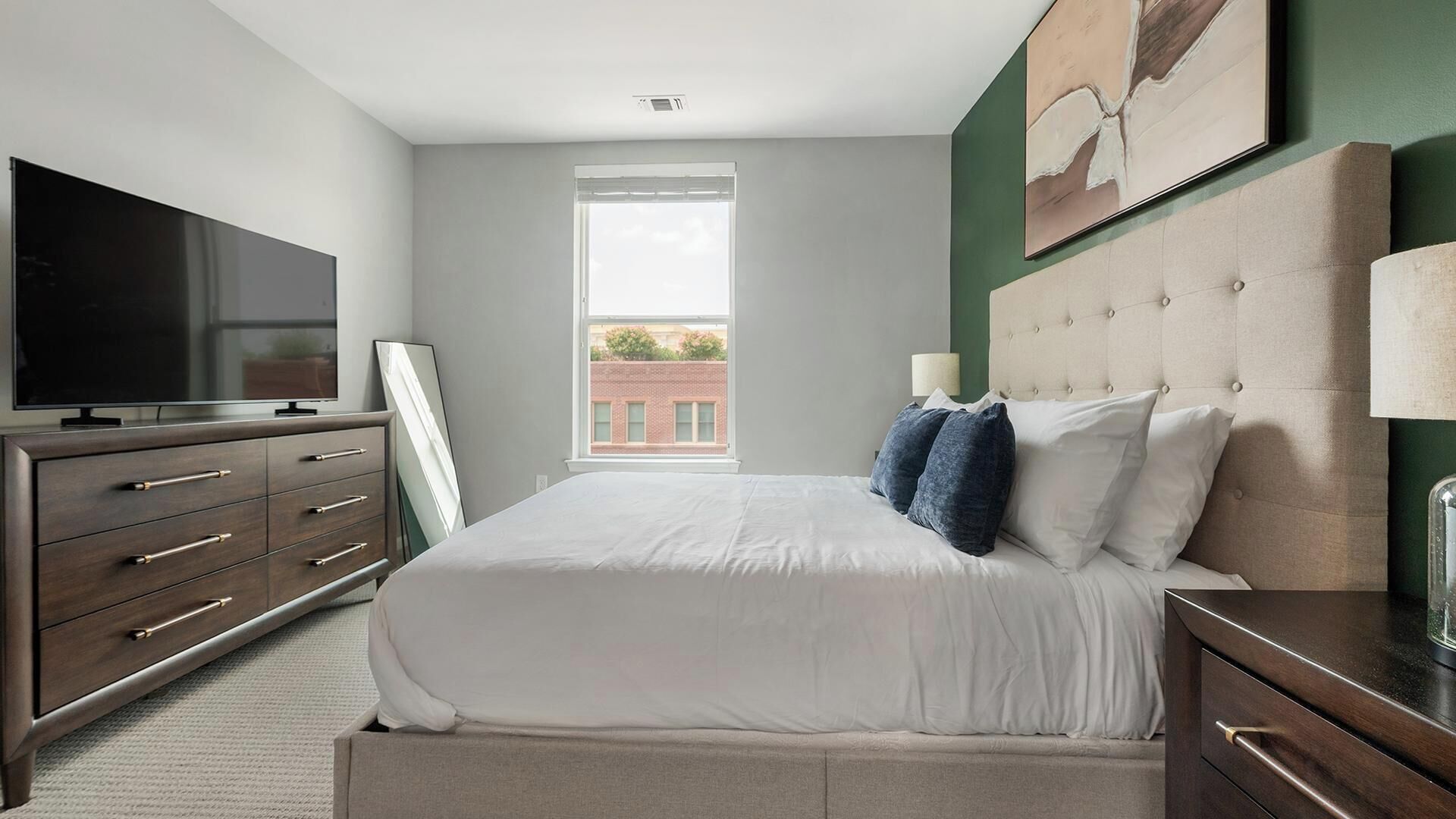 A bedroom with a large bed, a dresser, a television, and a window with a view of buildings.