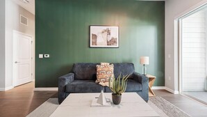 A living room with a dark green wall, a grey sofa, a patterned throw pillow, a potted plant, and a framed picture above the sofa. - Landing | Spectacular 1BD, Gym, Rooftop Lounge (Chevy Chase)