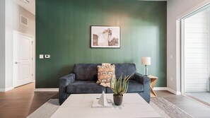 Ein Wohnzimmer mit dunkelgrüner Wand, einem grauen Sofa, einem gemusterten Sitzkissen, einer Topfpflanze und einem gerahmten Bild über dem Sofa.