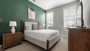 A bedroom with a bed, a nightstand, a lamp, a dresser, and a window with blinds. - Landing | Spectacular 1BD, Gym, Rooftop Lounge (Chevy Chase)