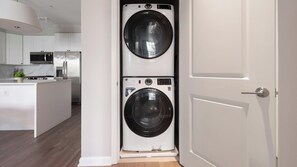 A laundry room with two stacked front-loading washing machines, a microwave, and a refrigerator in the adjacent kitchen area. - Landing | Modern 2BD, Gym, Rooftop Lounge (Chevy Chase)