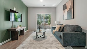 A modern living room with a flat-screen TV, a sofa, a coffee table, and a view through a glass door to an outdoor area. - Landing | Chic 1BD, Rooftop Lounge, Gym (Chevy Chase)