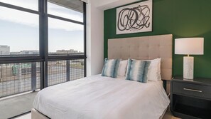 Quarto de hotel com uma cama grande, cabeceira, mesa de cabeceira, abajur e vista da cidade através de uma janela ampla.