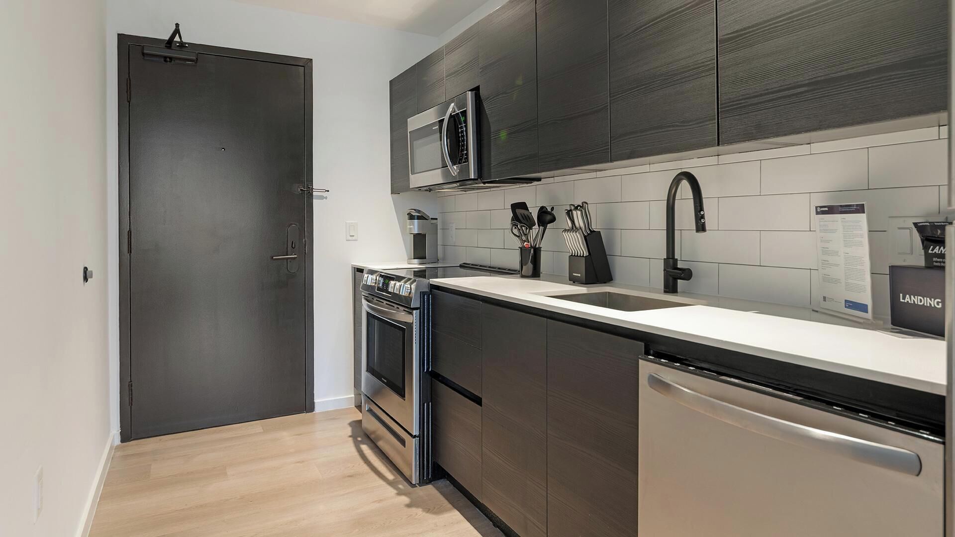 A modern kitchen with a built-in oven, microwave, and dishwasher, featuring dark cabinetry and a light countertop.