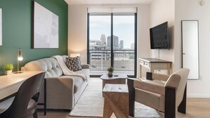 A modern living room with a sofa, coffee table, and chairs, a desk with a lamp, a TV mounted on the wall, and a large window with a city view. - Landing | Modern 1BD, Courtyard, Pool (Orlando)