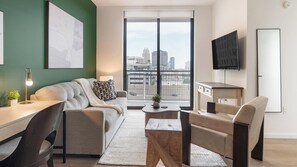 Ein modernes Wohnzimmer mit einer Couch, einem Couchtisch und Sesseln, einem Schreibtisch mit einer Lampe, einem an der Wand montierten Fernseher und einem großen Fenster mit Blick auf die Stadt.