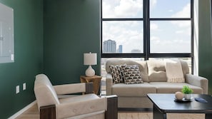 A modern living room with a green wall, a beige sofa, a white armchair, a wooden side table with a lamp, and a view of the city through large windows. - Landing | Beautiful 2BD, Pool, Rooftop Lounge (Orlando)