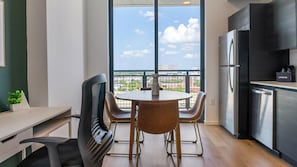 A modern kitchen with a stainless steel refrigerator, a dining area with a round table and chairs, and a large window offering a view of buildings and greenery. - Landing | Beautiful 2BD, Pool, Rooftop Lounge (Orlando)