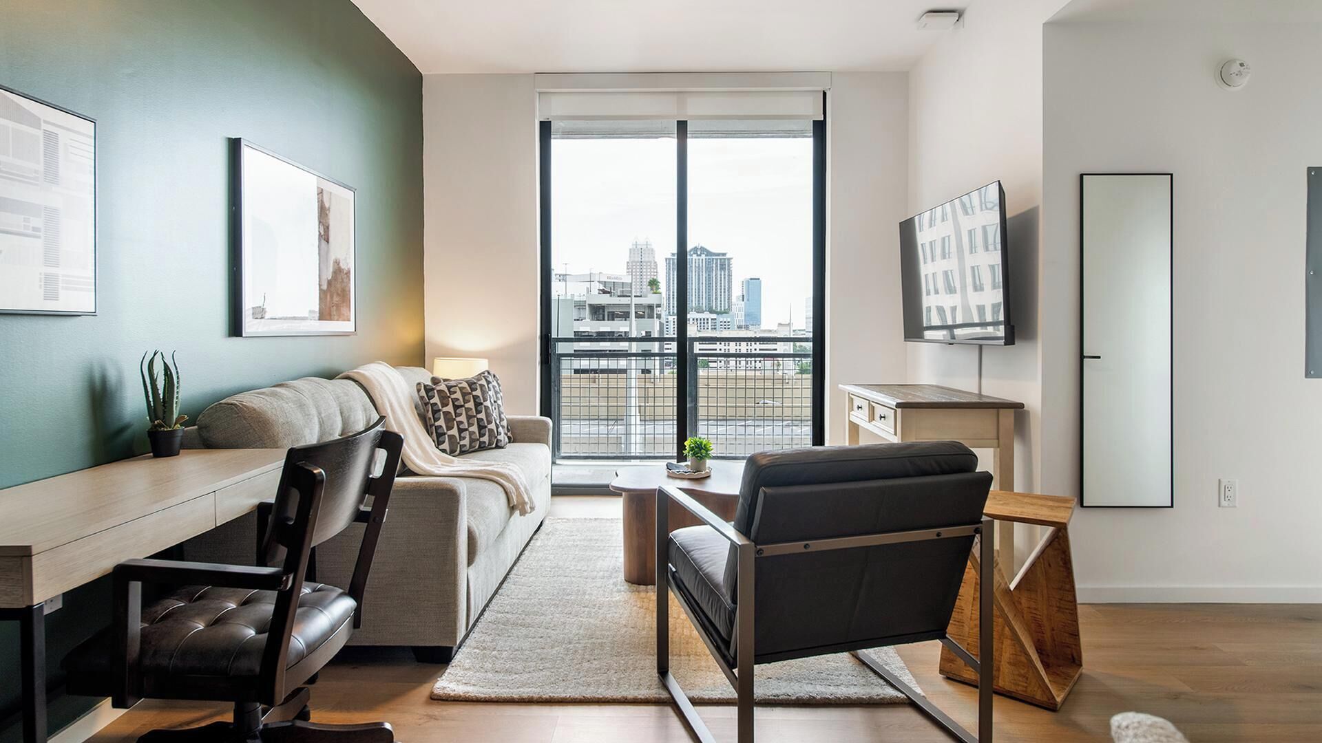 A modern living room with a sofa, a desk, a chair, a TV, and a large window with a city view.