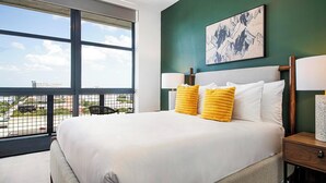 A hotel room with a large window, a bed with white linens, two yellow pillows, a headboard, and a nightstand with a lamp. - Landing | Stylish 2BD, Courtyard, Pool (Orlando)