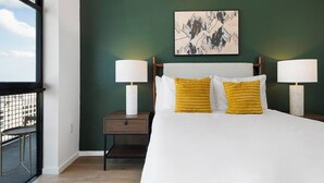 A hotel room with a large bed, two bedside lamps, a nightstand, a framed artwork on the wall, and a view through a window. - Landing | Stylish 2BD, Courtyard, Pool (Orlando)
