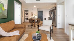 A modern living room with a sofa, coffee table, and a dining area with chairs. - Landing | Amazing 2BD, Pool, Gym (Orlando)