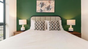 A bed with white bedding, two nightstands with lamps, and a green wall with a framed artwork above. - Landing | Amazing 2BD, Pool, Gym (Orlando)