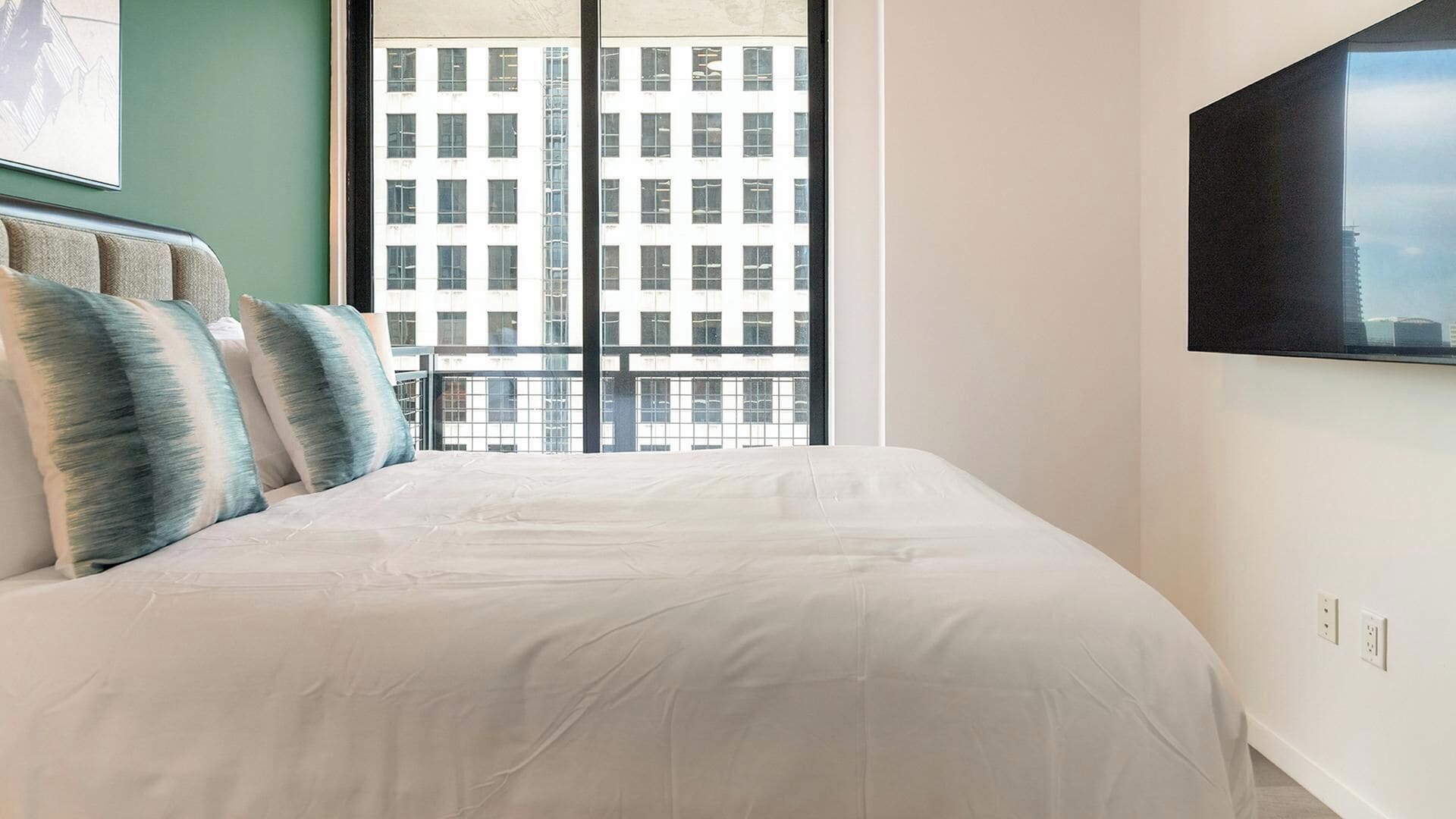 Ein Schlafzimmer mit einem großen Bett, einem Fenster mit Blick auf ein Gebäude und einem an der Wand befestigten Fernseher.