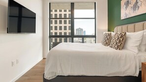 A bedroom with a large bed, a television mounted on the wall, and a view of a cityscape through a window. - Landing | Amazing 2BD, Pool, Gym (Orlando)