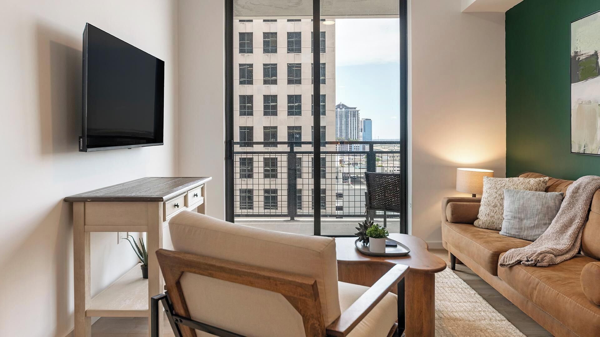A modern living room with a flat-screen TV mounted on the wall, a wooden desk, a comfortable sofa, and a large window offering a city view.