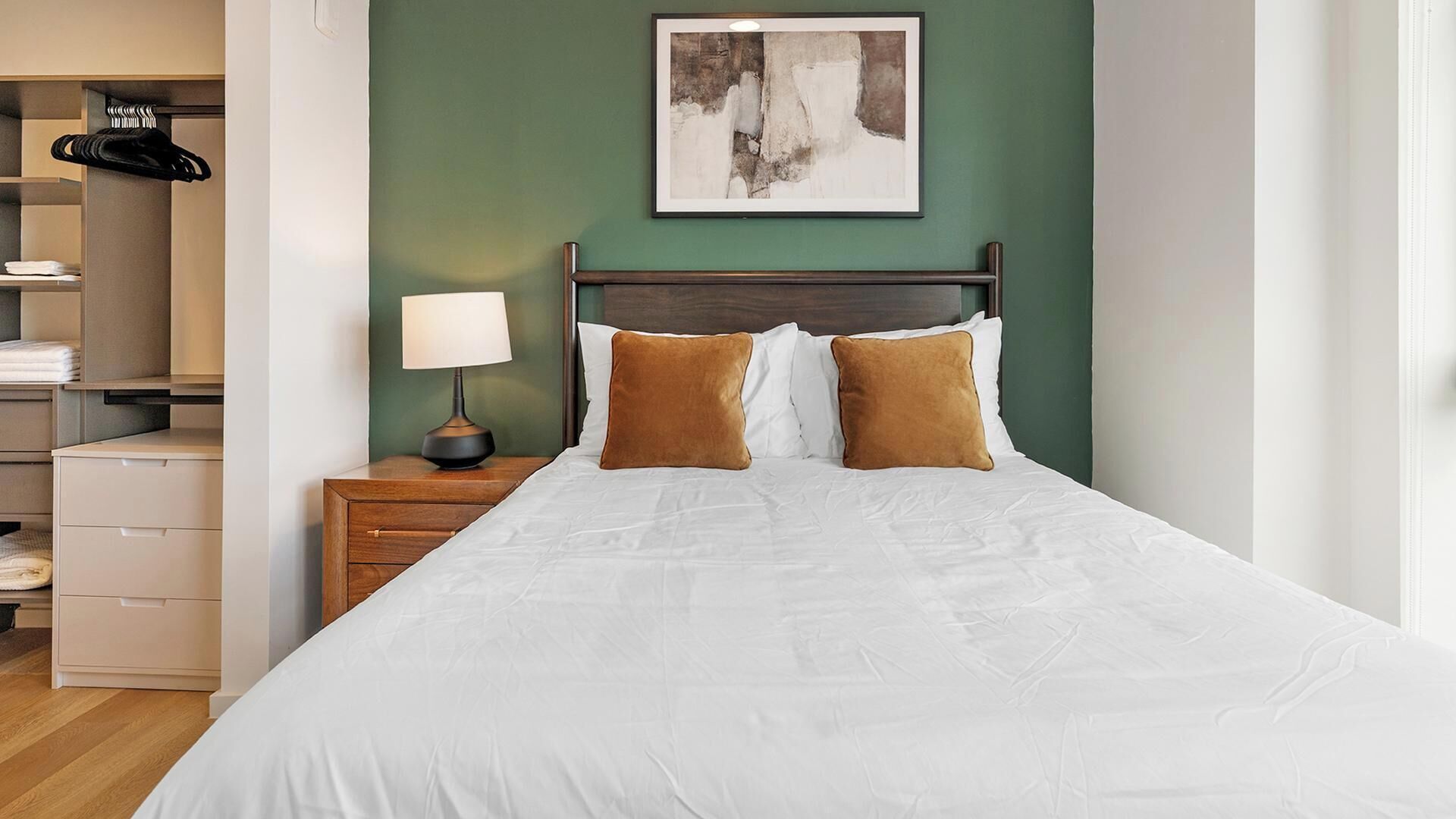 A neatly made bed with white bedding and two brown pillows, a bedside table with a lamp, a framed abstract painting on the wall, and a closet with hanging clothes.
