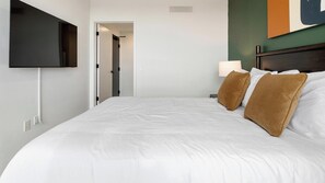 A hotel room with a bed, a flat-screen TV mounted on the wall, a bedside table with a lamp, and a door leading to another room. - Landing | Stunning 2BD, Gym, Rooftop Lounge (Orlando)