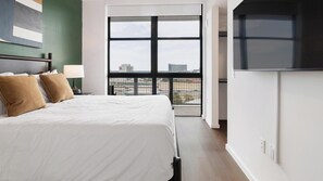 A hotel room with a large bed, a nightstand, a lamp, and a view of the city through a large window. - Landing | Stunning 2BD, Gym, Rooftop Lounge (Orlando)