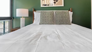 A neatly made bed with white linens and two patterned pillows, a bedside lamp, and a wall art piece above the bed. - Landing | Chic Studio, Rooftop Lounge, Courtyard (Orlando)