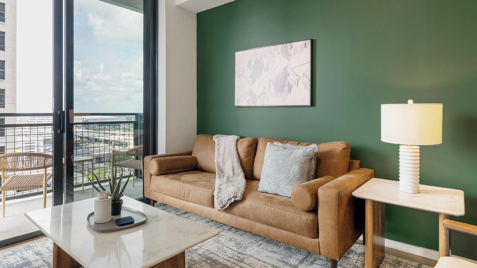 A modern living room with a leather sofa, a coffee table, a lamp, and a view of the city through large windows.